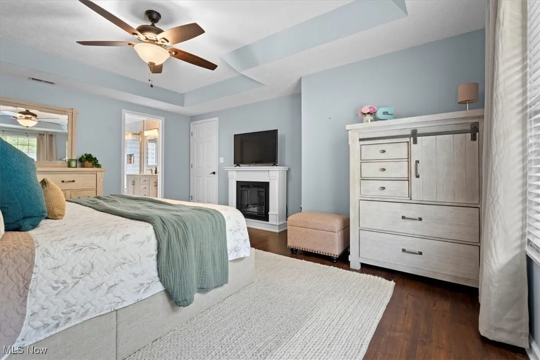 Bedroom featuring a raised ceiling, a glass covered fireplace, dark wood-style flooring, a ceiling fan, and ensuite bathroom
