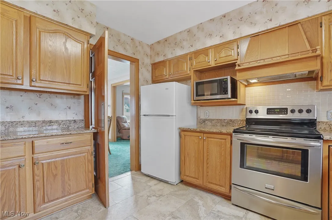 Kitchen with stainless steel appliances, decorative backsplash, wallpapered walls, premium range hood, and light stone countertops