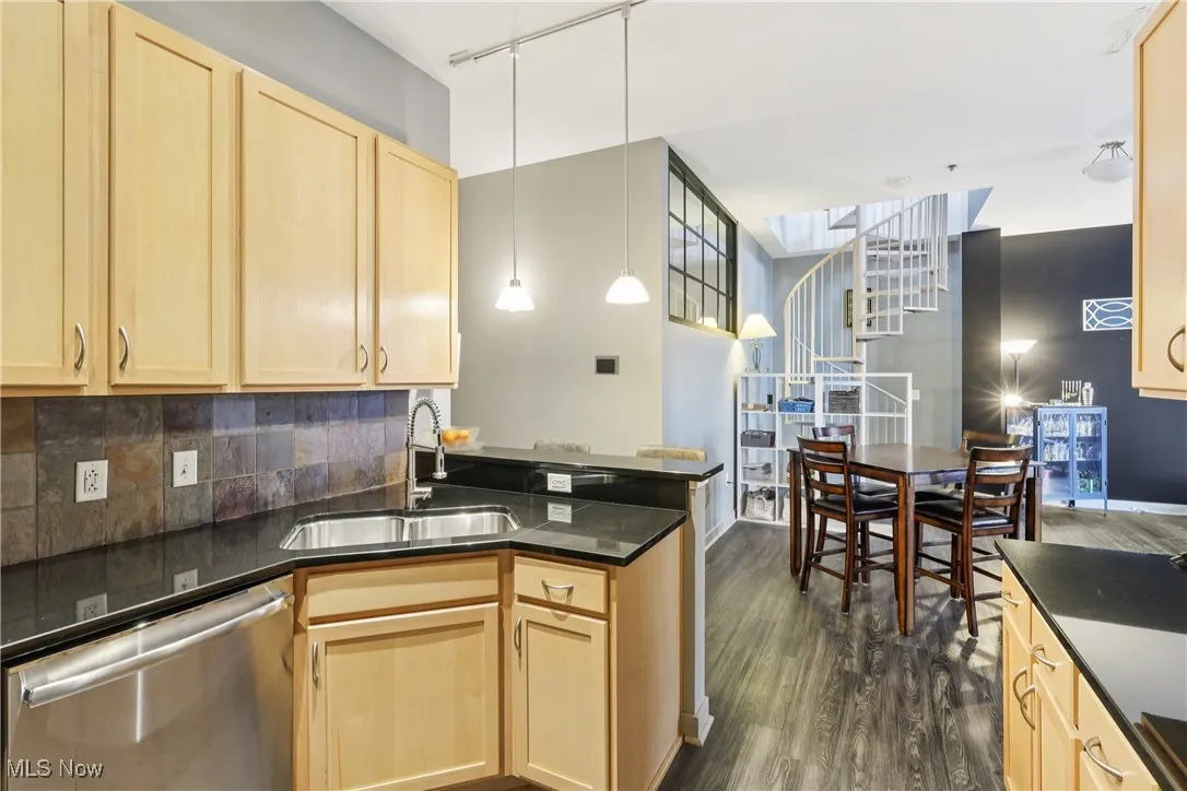 Kitchen featuring light brown cabinetry, stainless steel dishwasher, dark wood-style flooring, hanging light fixtures, and decorative backsplash