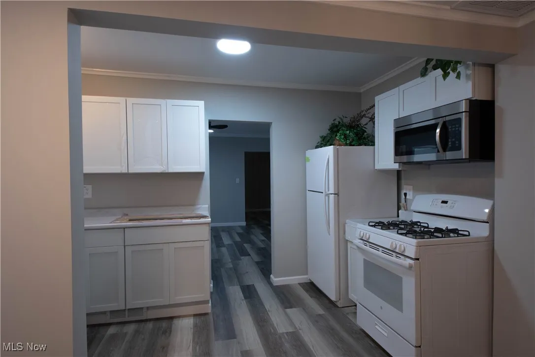 Kitchen with crown molding, white appliances, white cabinetry, light countertops, and dark wood-style flooring