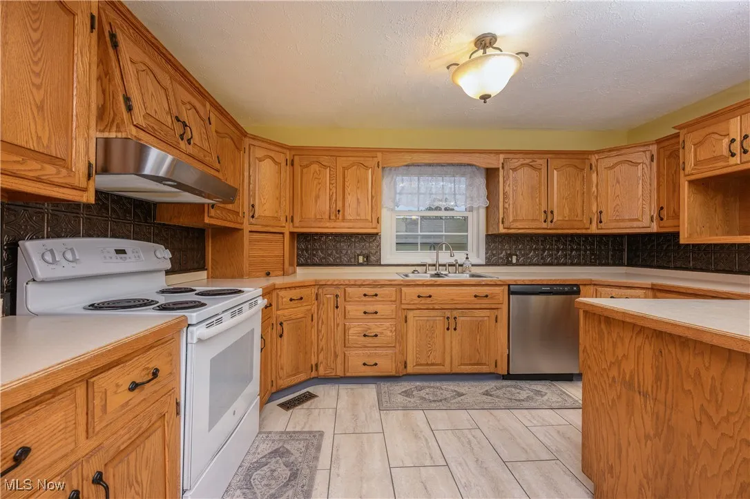 Kitchen with white range with electric stovetop, brown cabinetry, under cabinet range hood, dishwasher, and backsplash