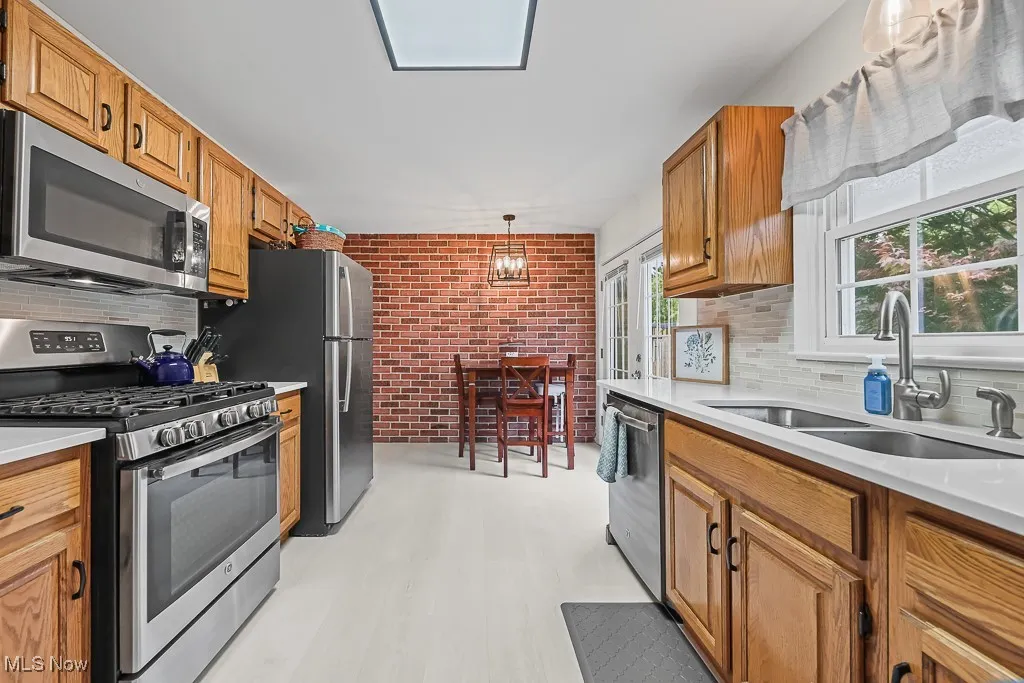 Kitchen featuring stainless steel appliances, brick wall, decorative backsplash, brown cabinetry, and decorative light fixtures