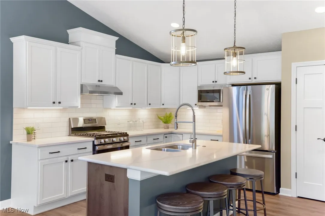 Kitchen featuring decorative light fixtures, a kitchen island with sink, appliances with stainless steel finishes, vaulted ceiling, and white cabinets