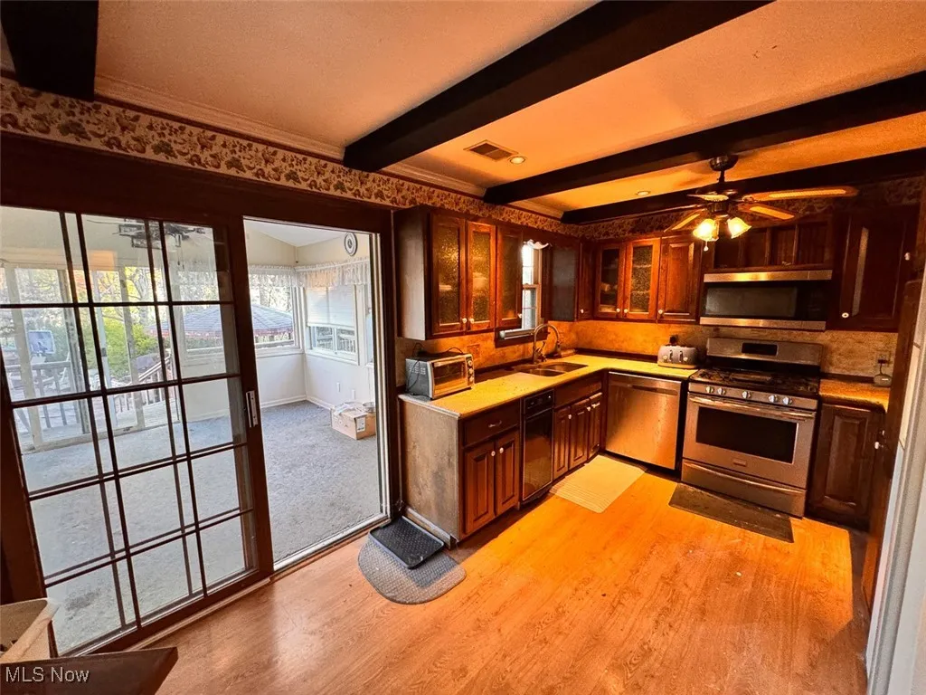 Kitchen with stainless steel appliances, glass insert cabinets, light wood-type flooring, beam ceiling, and a ceiling fan