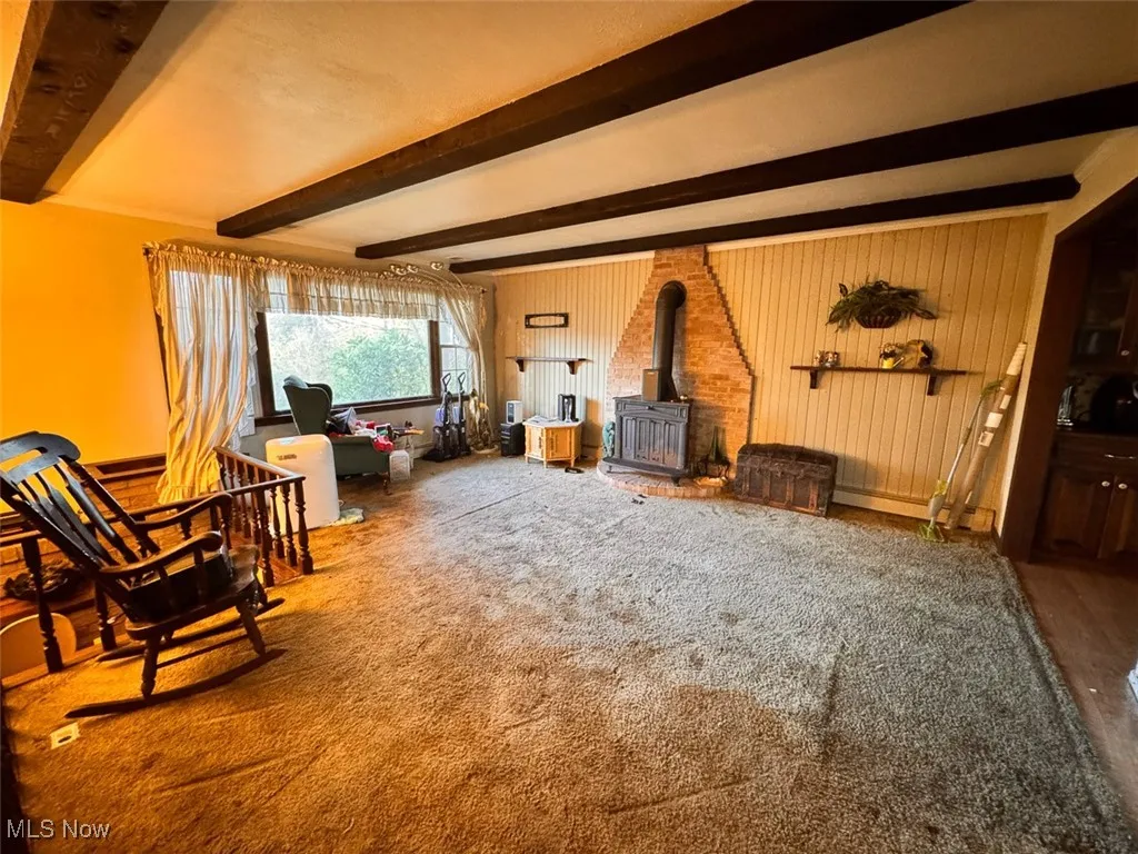 Living room featuring a wood stove, beamed ceiling, carpet, and wooden walls