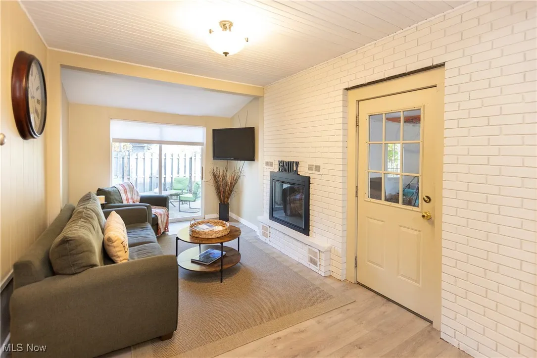 Living room featuring brick wall, wood finished floors, a fireplace, and beamed ceiling leading to the outdoor patio