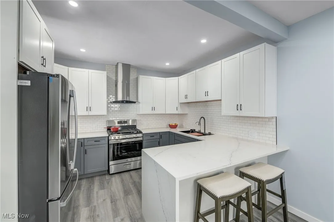Kitchen featuring gray cabinets, a peninsula, stainless steel appliances, wall chimney exhaust hood, and a sink