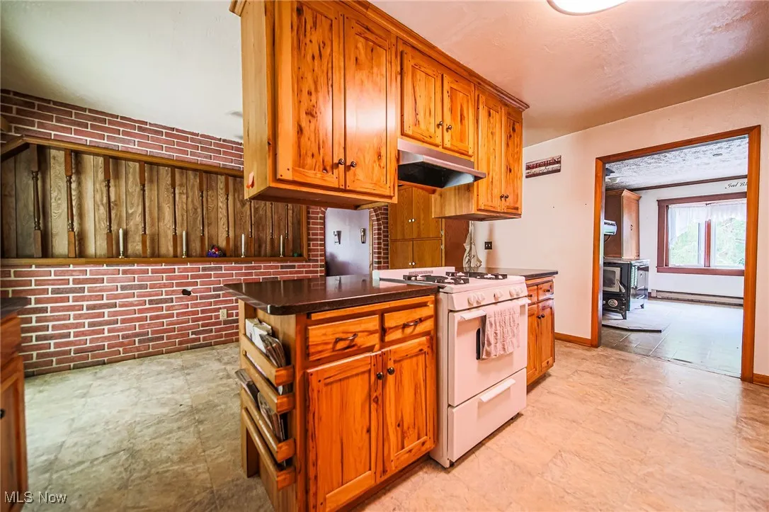 Kitchen with white gas range oven, dark countertops, brick wall, brown cabinets, and under cabinet range hood