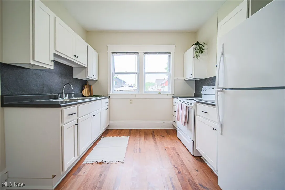 Kitchen with white appliances, a sink, dark countertops, white cabinetry, and light wood finished floors