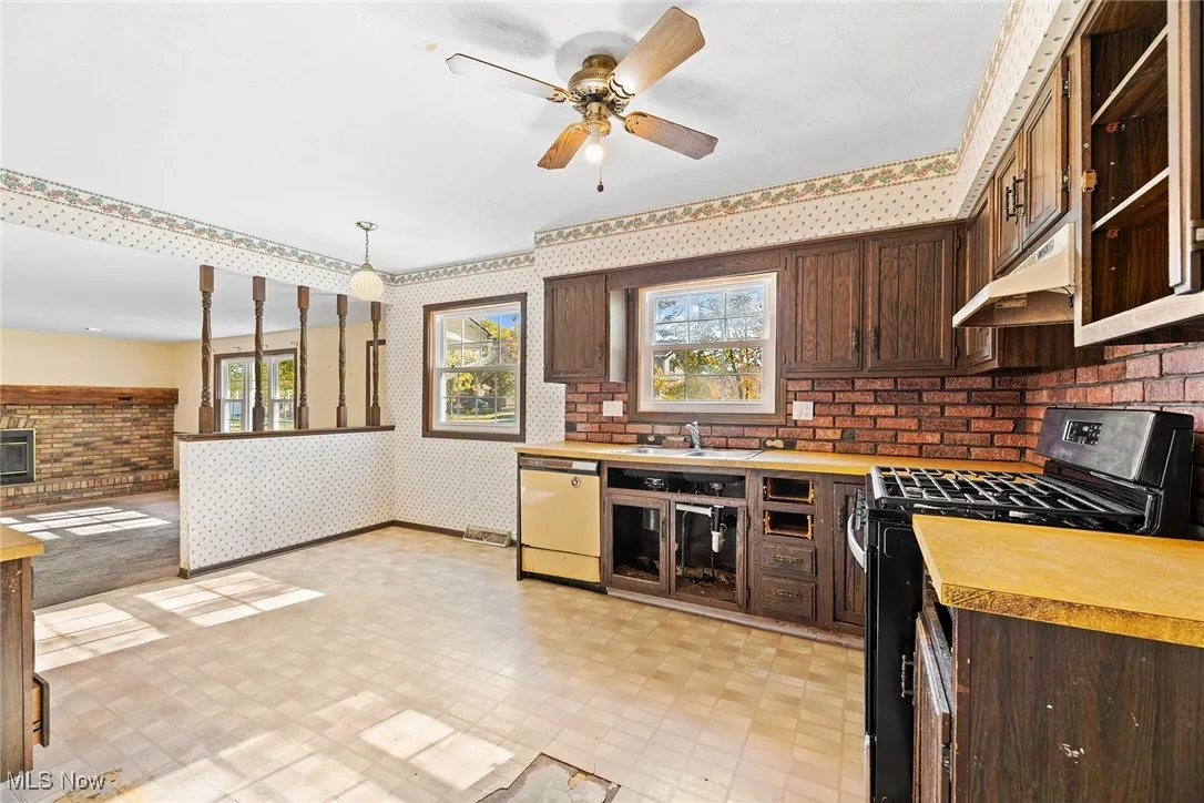 Kitchen with dark brown cabinets, black gas stove, wallpapered walls, dishwashing machine, and a ceiling fan
