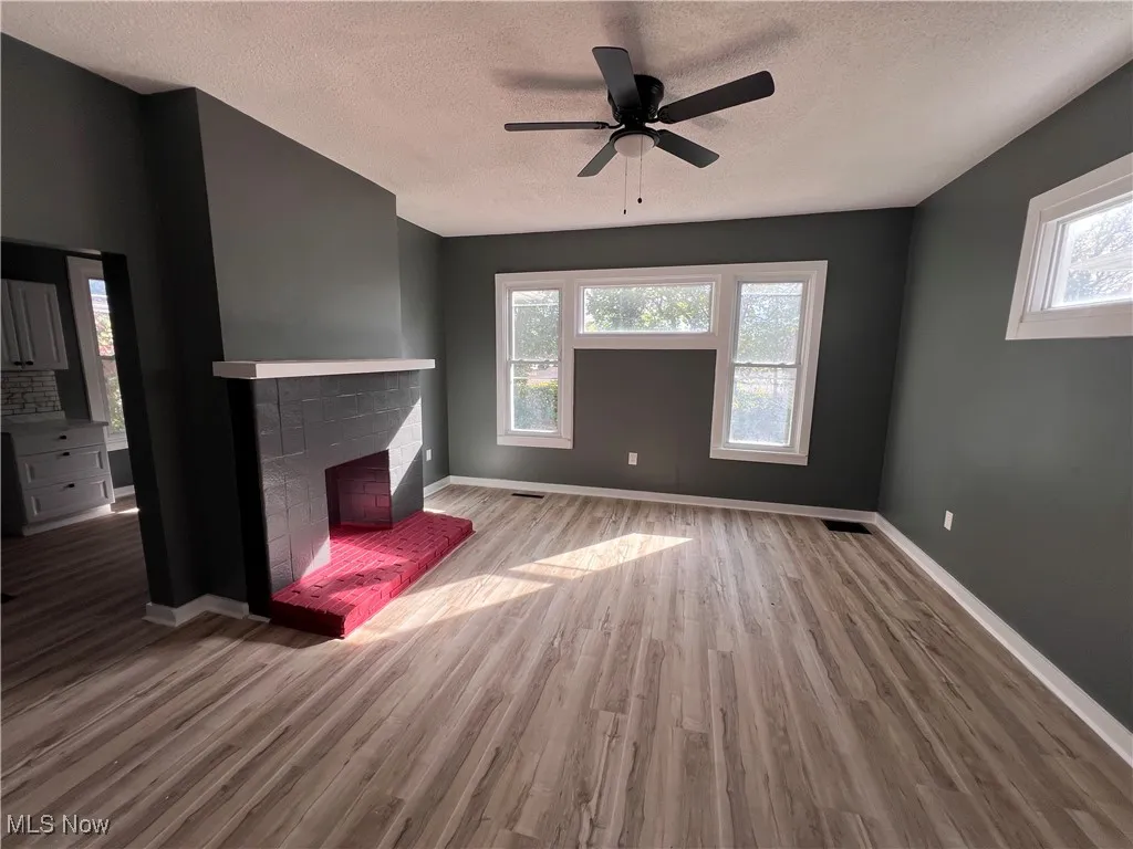 Unfurnished living room featuring a brick fireplace, wood finished floors, a ceiling fan, and a textured ceiling