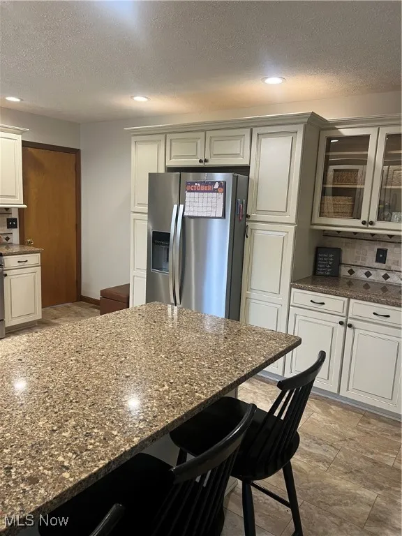Kitchen with backsplash, stainless steel refrigerator with ice dispenser, a textured ceiling, a kitchen bar, and dark stone countertops