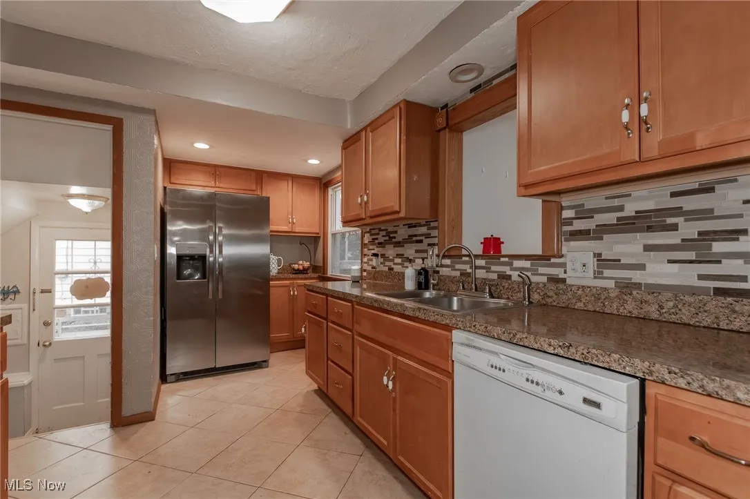 Kitchen with stainless steel refrigerator with ice dispenser, white dishwasher, decorative backsplash, light tile patterned floors, and dark countertops