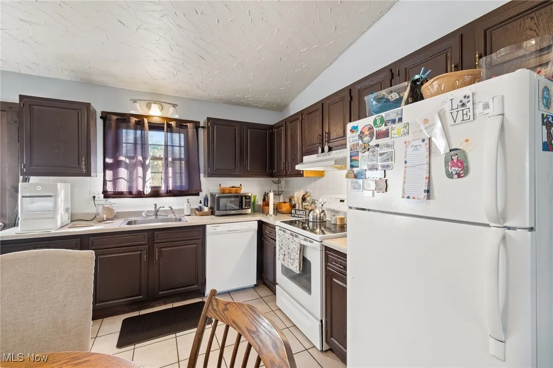 Kitchen featuring white appliances, light tile patterned flooring, light countertops, dark brown cabinetry, and under cabinet range hood