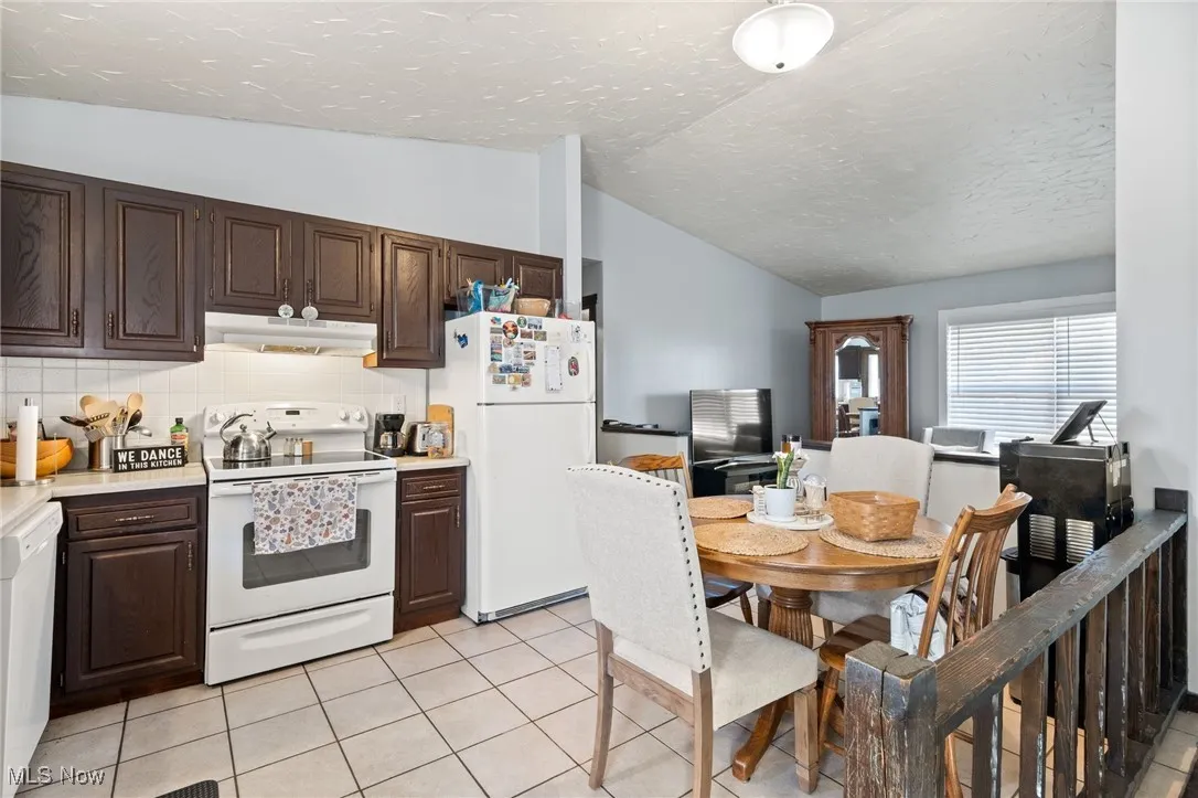 Kitchen featuring white appliances, light countertops, dark brown cabinetry, light tile patterned floors, and vaulted ceiling