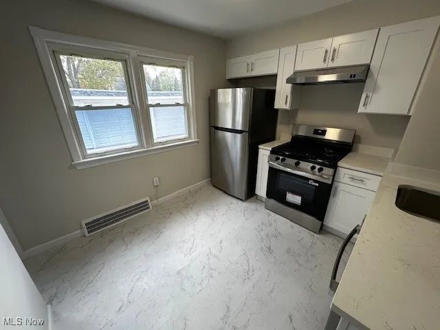 Kitchen with white cabinets, stainless steel range with gas cooktop, light stone counters, under cabinet range hood, and light marble finish flooring