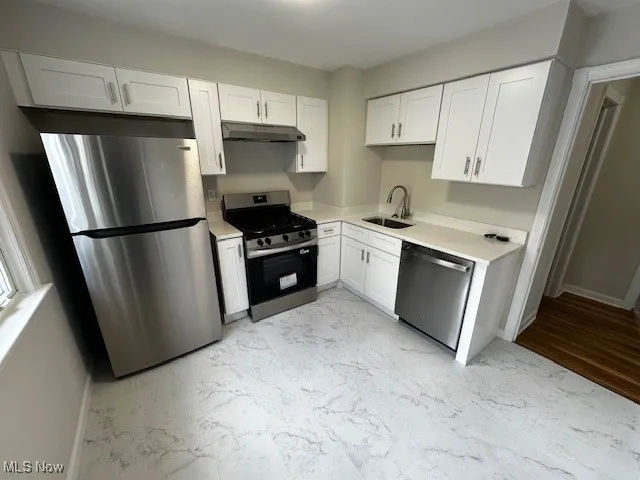 Kitchen featuring appliances with stainless steel finishes, white cabinets, light marble finish flooring, under cabinet range hood, and light stone counters