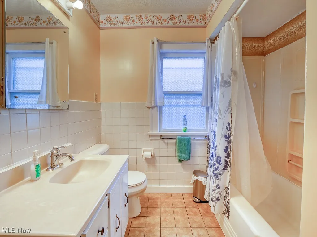 Bathroom with light tile patterned floors, vanity, shower / bathtub combination with curtain, tile walls, and wainscoting