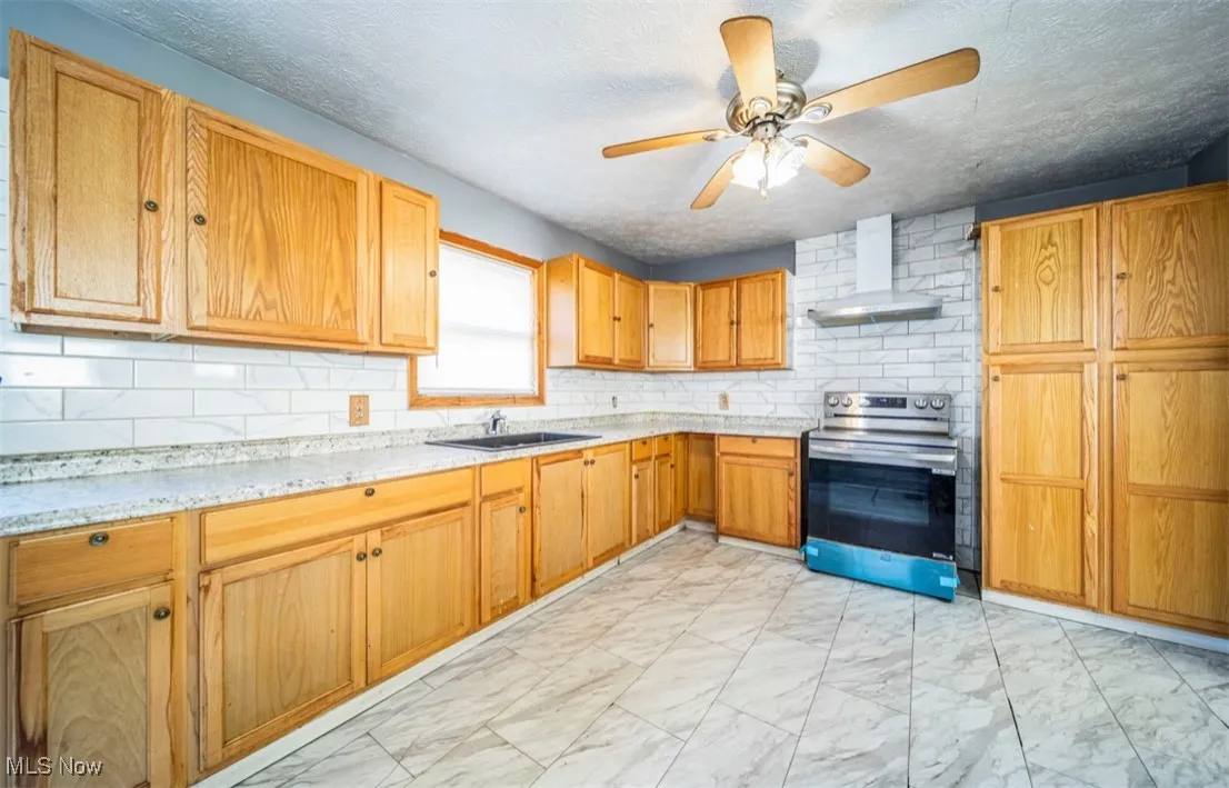 Kitchen with light marble finish floors, stainless steel range with electric cooktop, wall chimney exhaust hood, a ceiling fan, and backsplash