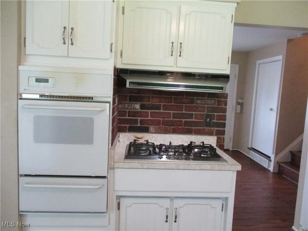 Kitchen with dark hardwood / wood-style floors, stainless steel gas stovetop, range hood, white cabinets + wall oven