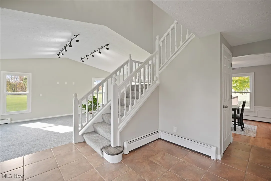Staircase with tile patterned floors, rail lighting, a textured ceiling, a baseboard radiator, and vaulted ceiling