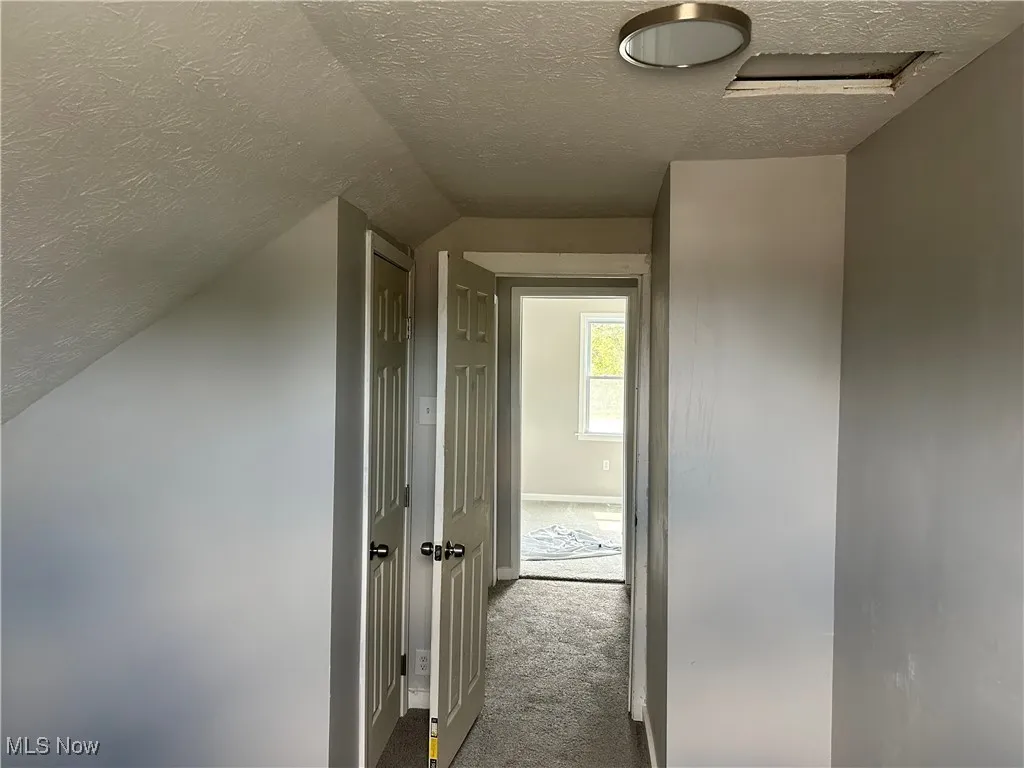 Hallway featuring a textured ceiling, dark carpet, vaulted ceiling, and attic access