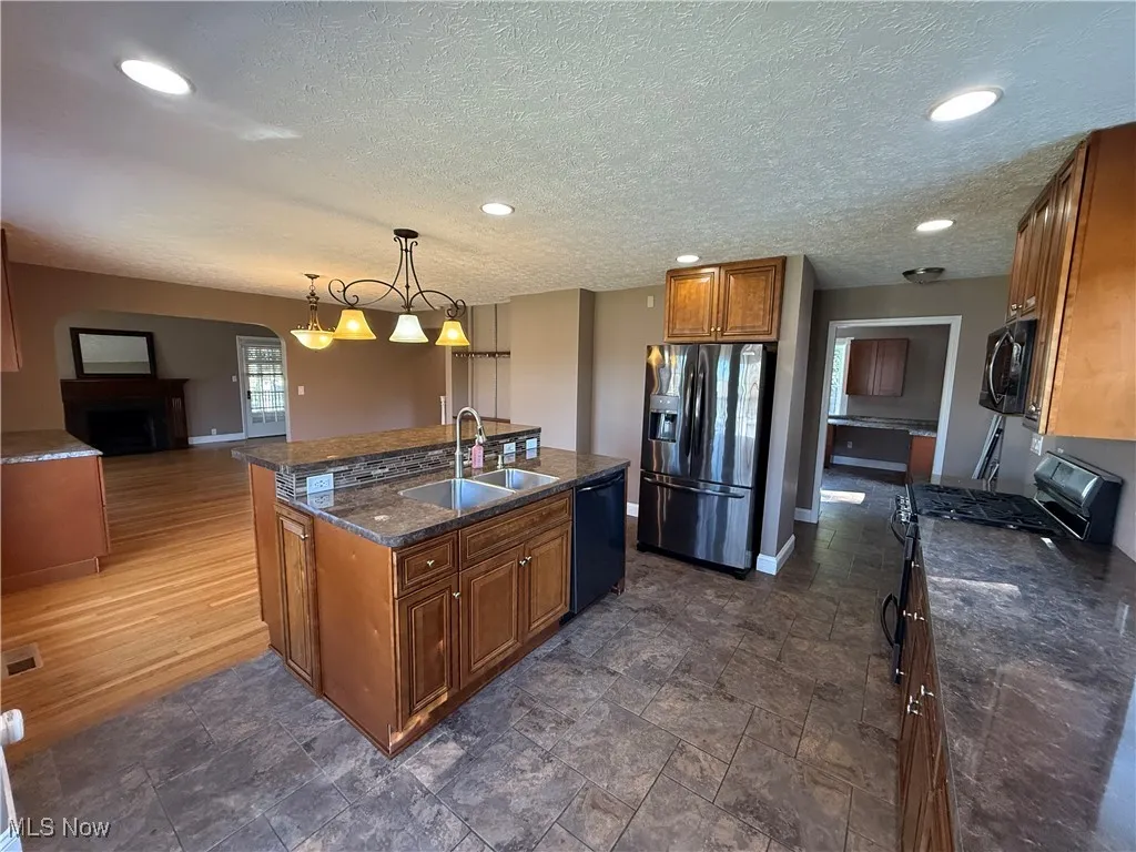 Kitchen with brown cabinets, black appliances, hanging light fixtures, a center island with sink, and arched walkways