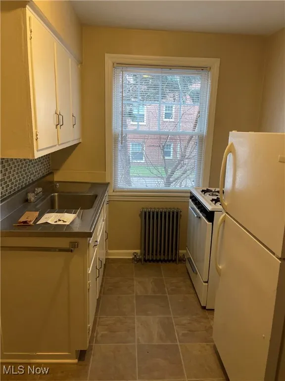 Kitchen featuring white appliances, radiator heating unit, white cabinetry, dark countertops, and tasteful backsplash