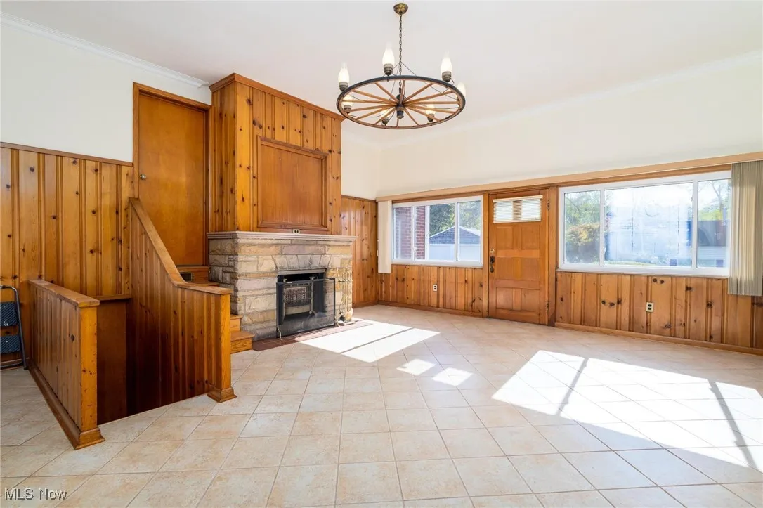 Unfurnished living room featuring wooden walls, crown molding, a stone fireplace, light tile patterned floors, and a chandelier