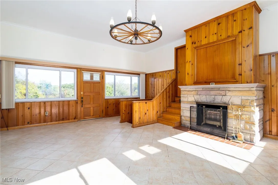 Unfurnished living room featuring ornamental molding, wooden walls, a chandelier, a stone fireplace, and stairs