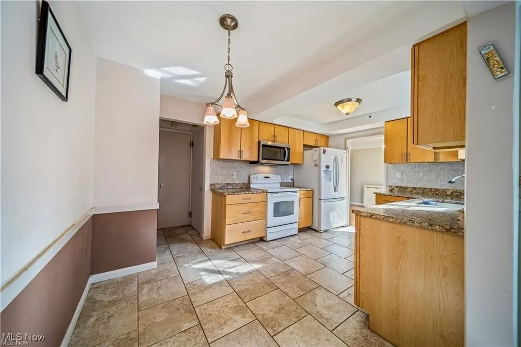Kitchen with white appliances, hanging light fixtures, light tile patterned flooring, decorative backsplash, and dark stone countertops