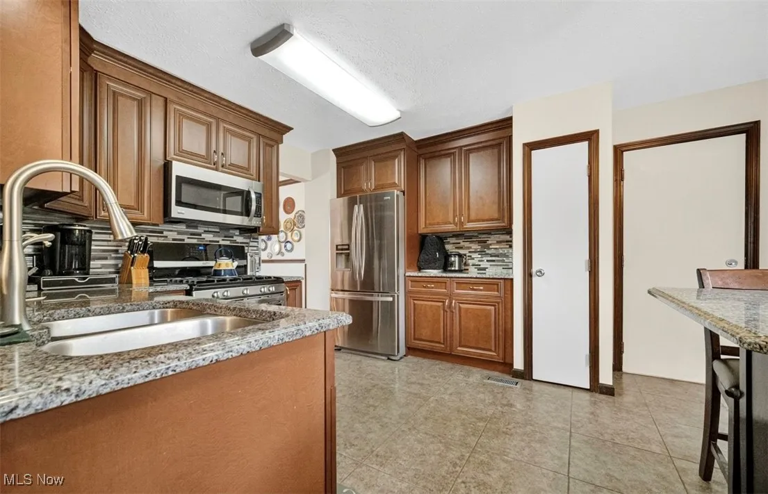 Kitchen with light stone countertops, appliances with stainless steel finishes, tasteful backsplash, light tile patterned floors, and brown cabinetry