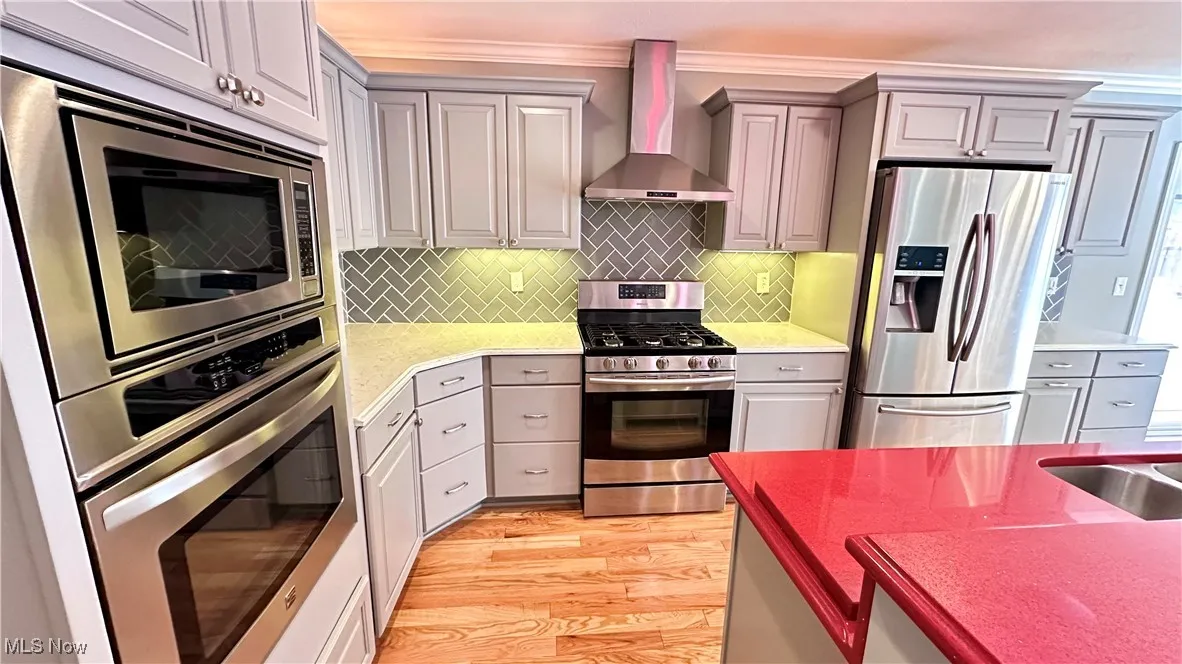 Kitchen featuring stainless steel appliances, wall chimney exhaust hood, gray cabinetry, tasteful backsplash, and light wood-style floors