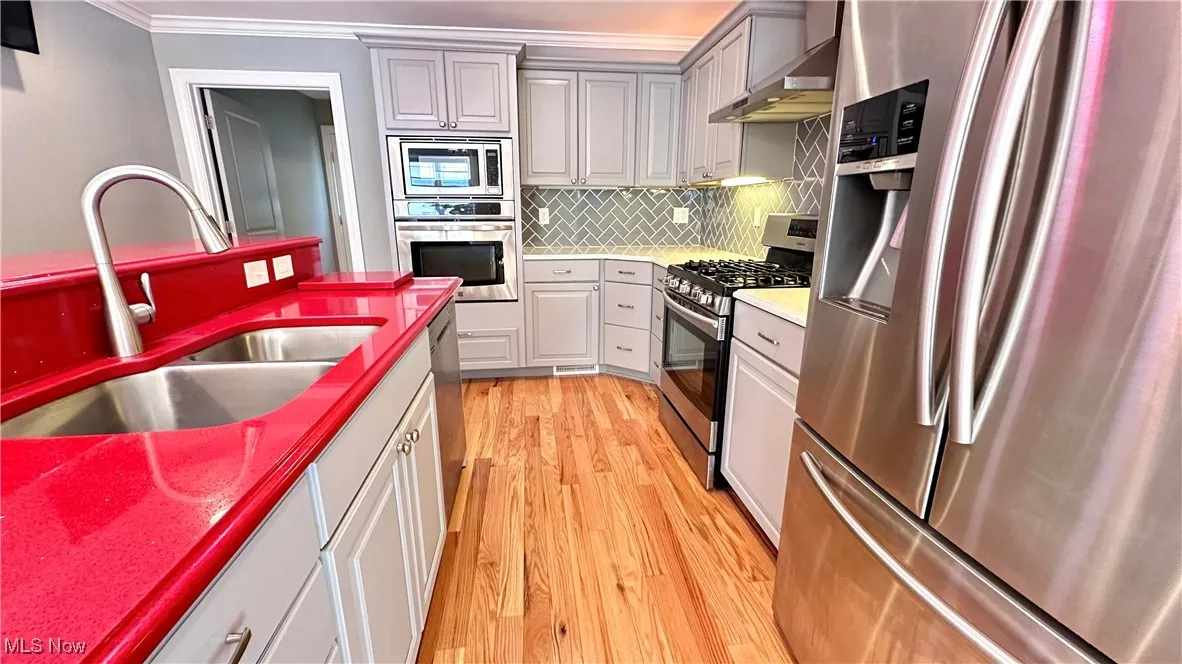 Kitchen featuring appliances with stainless steel finishes, wall chimney range hood, tasteful backsplash, light wood-style floors, and crown molding