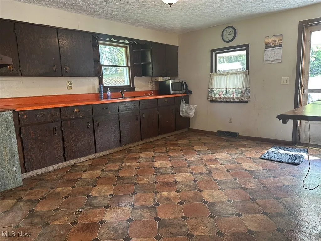 Kitchen featuring a textured ceiling, light countertops, dark brown cabinets, stainless steel microwave, and open shelves