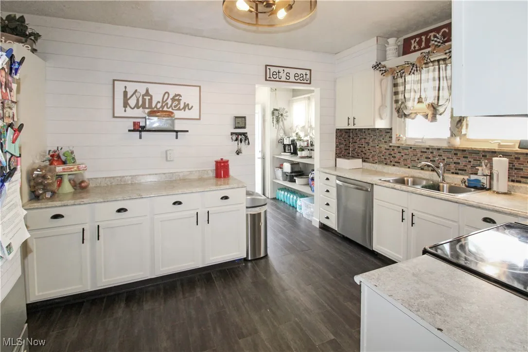 Kitchen with white cabinets, light countertops, dark wood finished floors, stainless steel dishwasher, and wooden walls