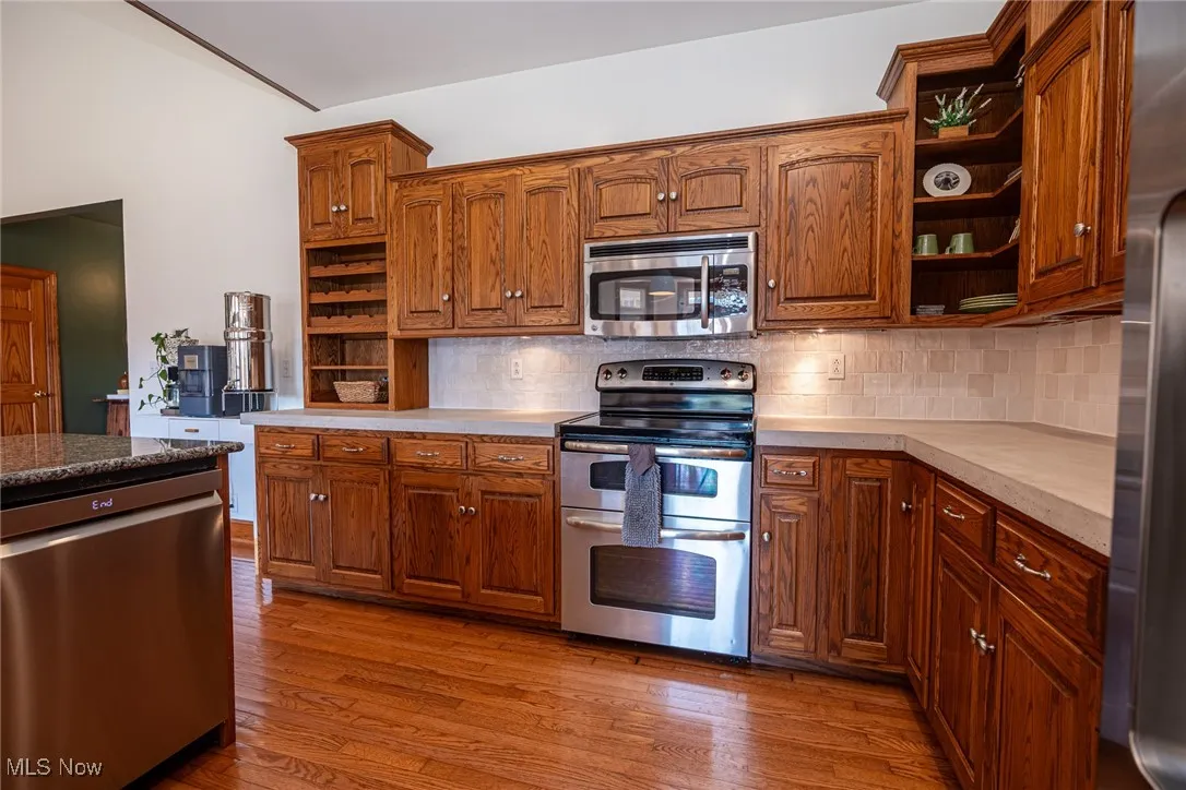 Kitchen with open shelves, appliances with stainless steel finishes, dark wood-style floors, decorative backsplash, and vaulted ceiling