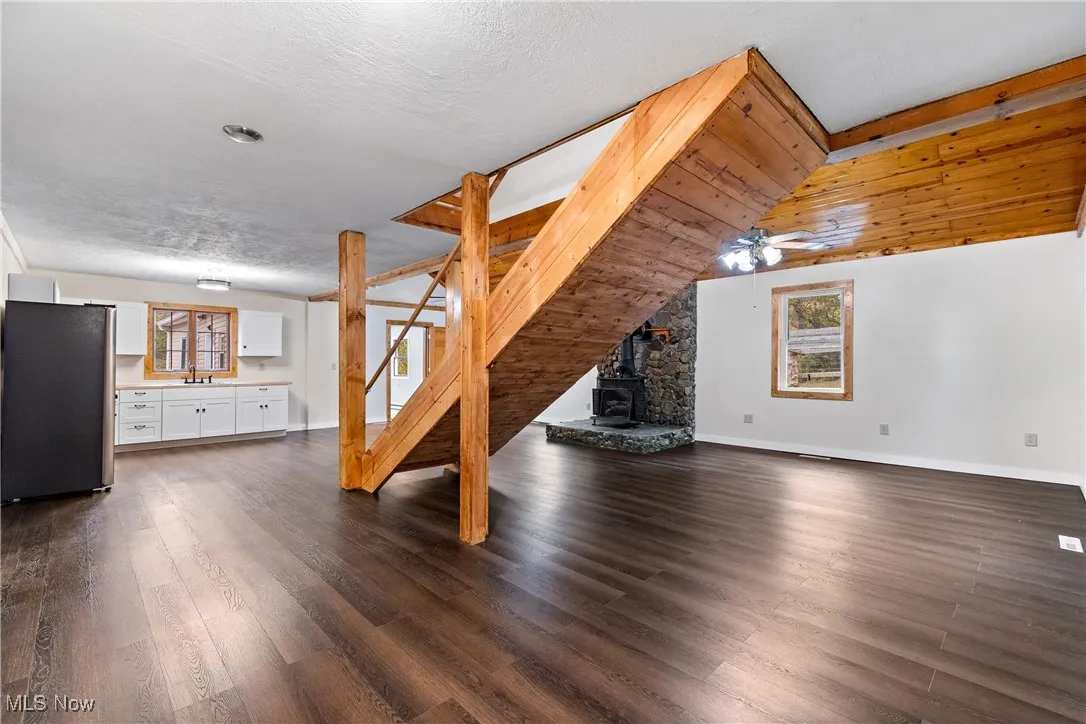 Unfurnished living room featuring dark wood-style floors, a wood stove, stairs, a ceiling fan, and a textured ceiling
