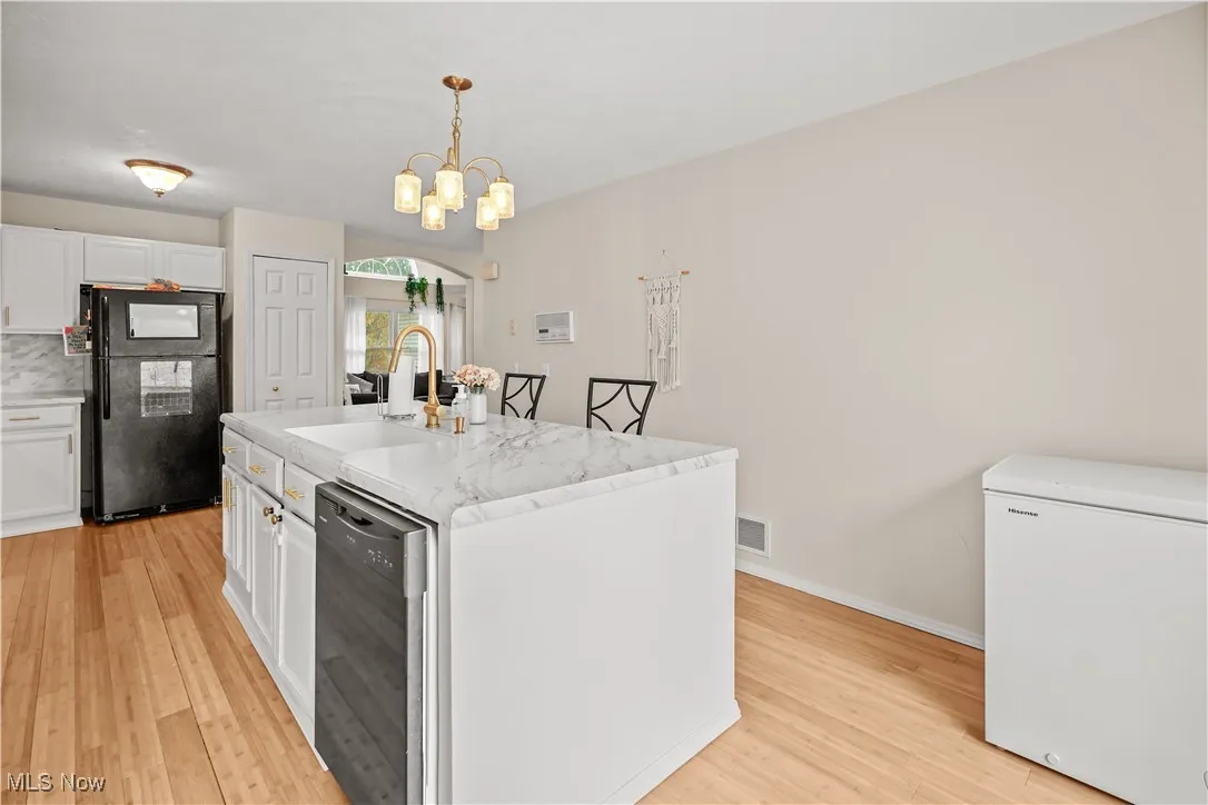 Kitchen featuring black appliances, white cabinets, light wood-type flooring, decorative light fixtures, and a center island with sink