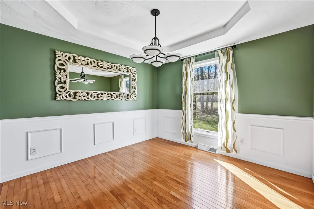 Unfurnished dining area with a raised ceiling, a wainscoted wall, a chandelier, wood-type flooring, and a decorative wall