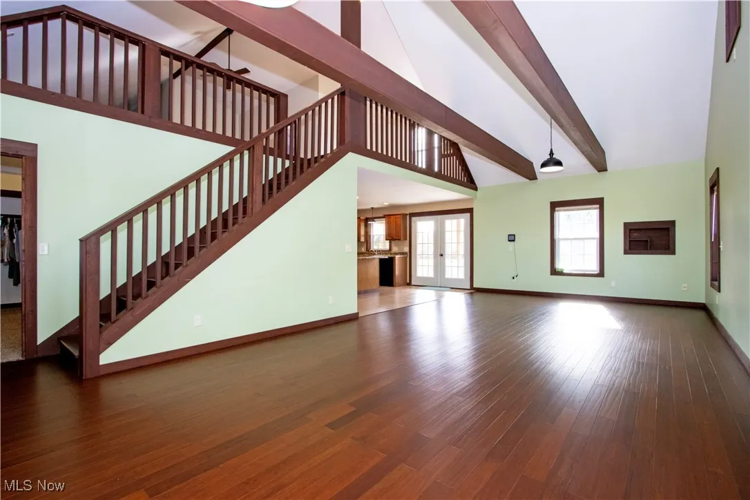 Unfurnished living room featuring high vaulted ceiling, dark wood finished floors, french doors, beamed ceiling, and stairs