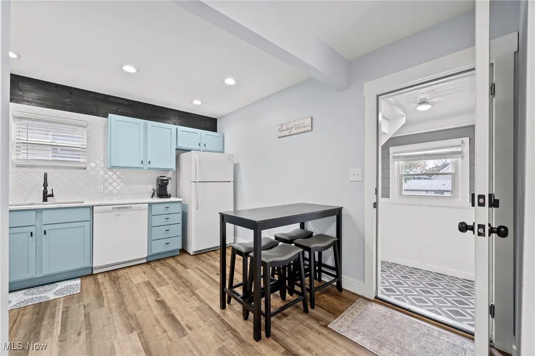 Kitchen featuring blue cabinets, white appliances, backsplash, light wood-style flooring, and beam ceiling