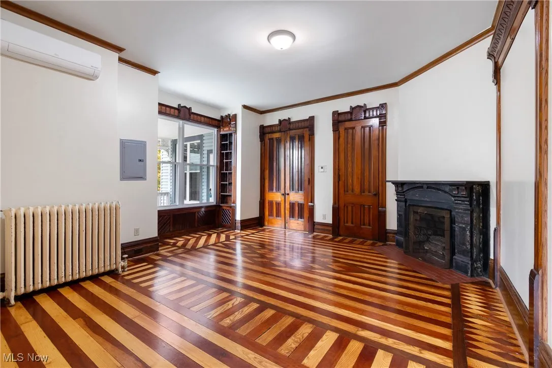 Unfurnished living room with crown molding, radiator, hardwood / wood-style flooring, a fireplace with flush hearth, and an AC wall unit