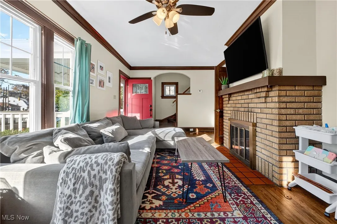 Living room with crown molding, a ceiling fan, dark wood-type flooring, arched walkways, and a brick fireplace