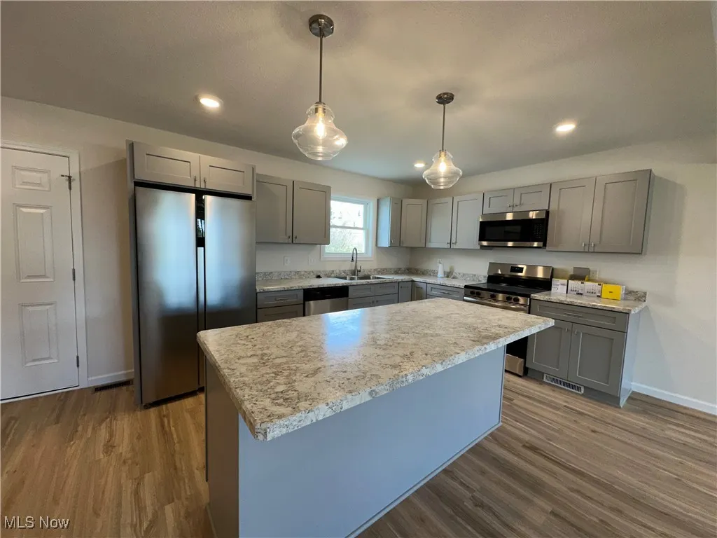Kitchen with gray cabinets, appliances with stainless steel finishes, hanging light fixtures, a kitchen island, and wood-style flooring