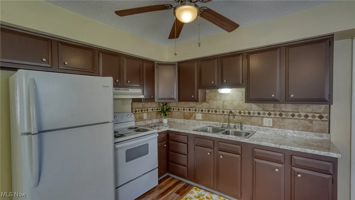 Kitchen with white appliances, a textured ceiling, backsplash, dark brown cabinetry, and under cabinet range hood