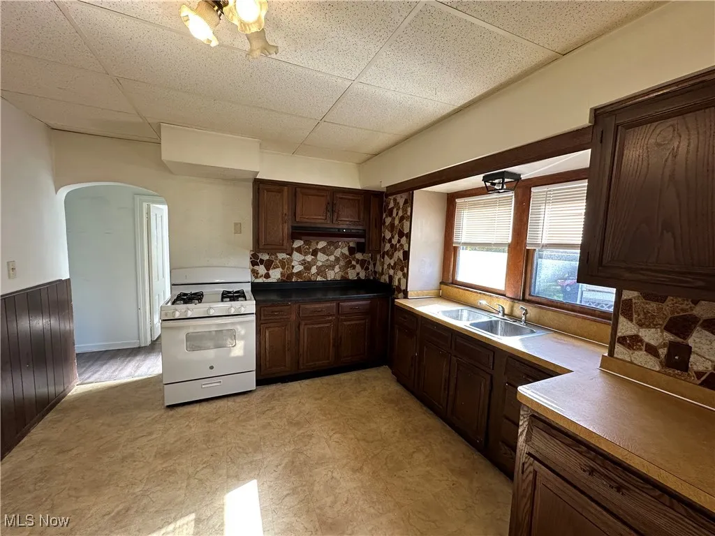 Kitchen with white range with gas stovetop, a paneled ceiling, arched walkways, dark brown cabinets, and tasteful backsplash