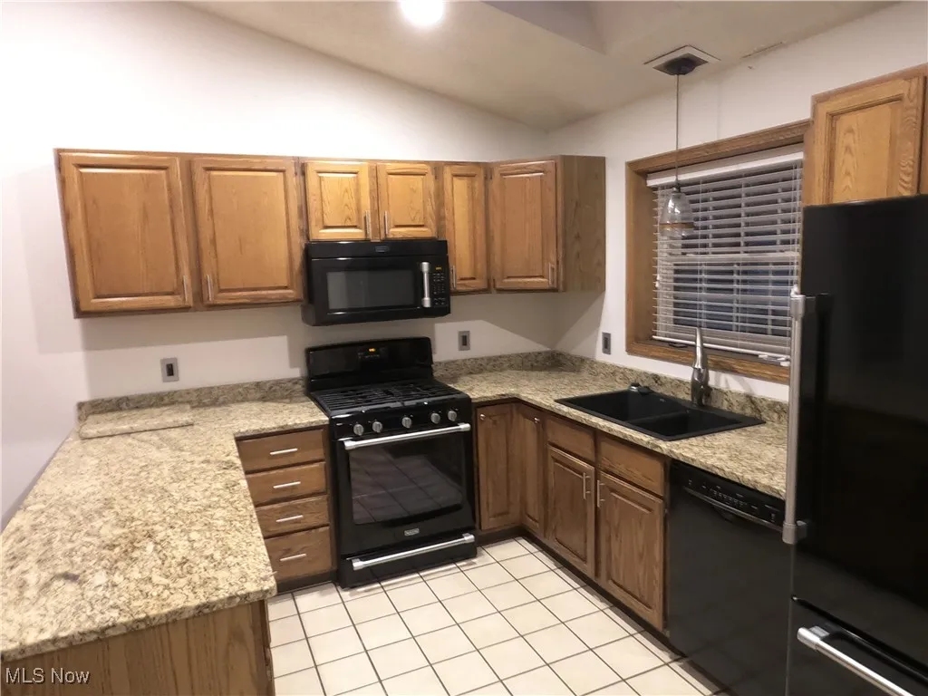 Kitchen with black appliances, light tile patterned flooring, lofted ceiling, light stone countertops, and decorative light fixtures