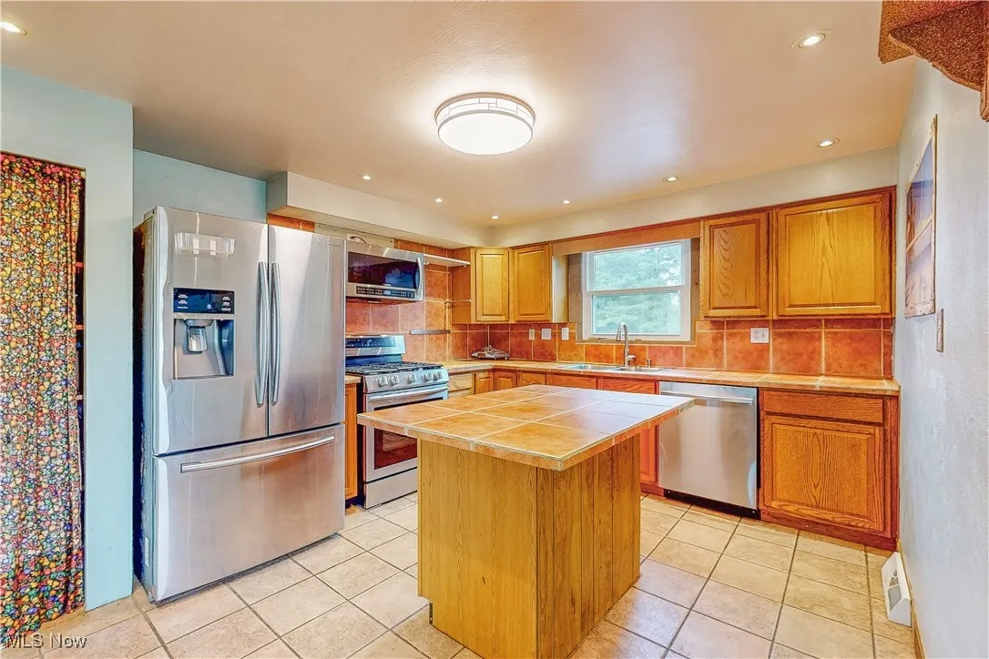 Kitchen with appliances with stainless steel finishes, brown cabinets, a kitchen island, backsplash, and light tile patterned floors