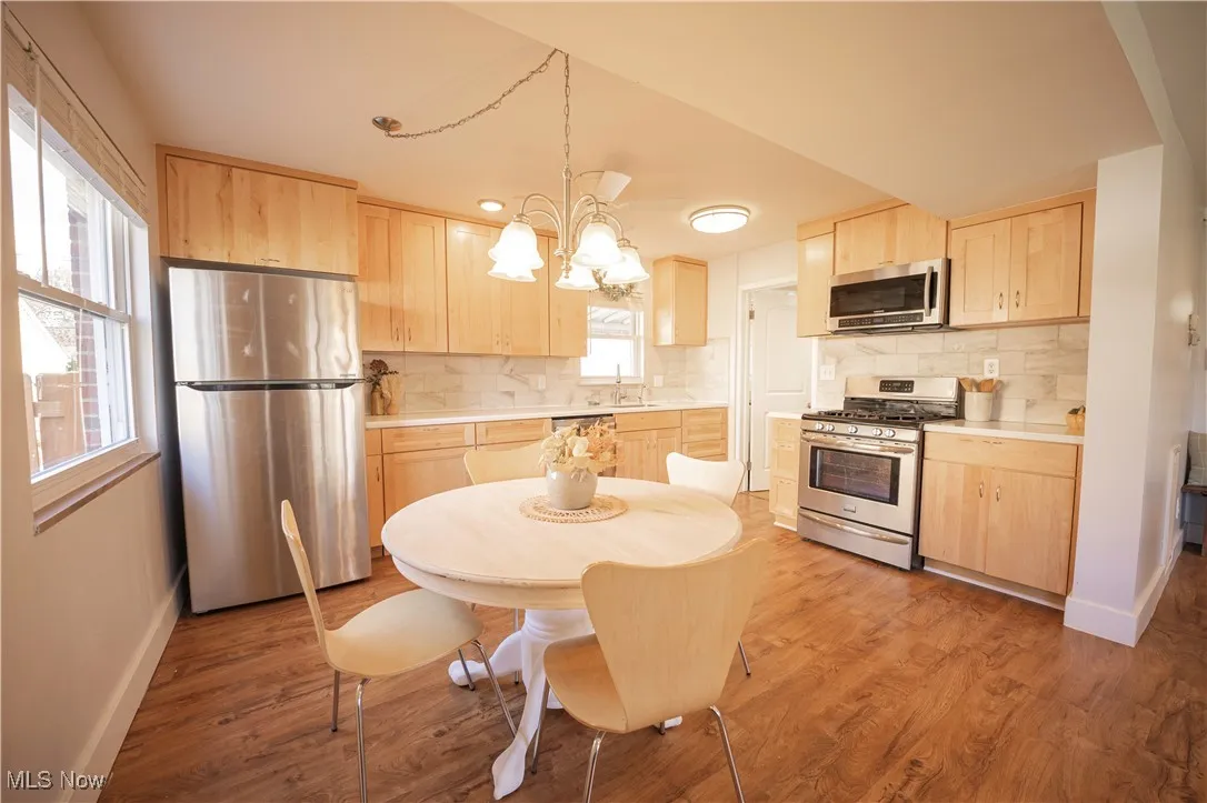 Kitchen with light brown cabinetry, appliances with stainless steel finishes, backsplash, and hanging light fixtures